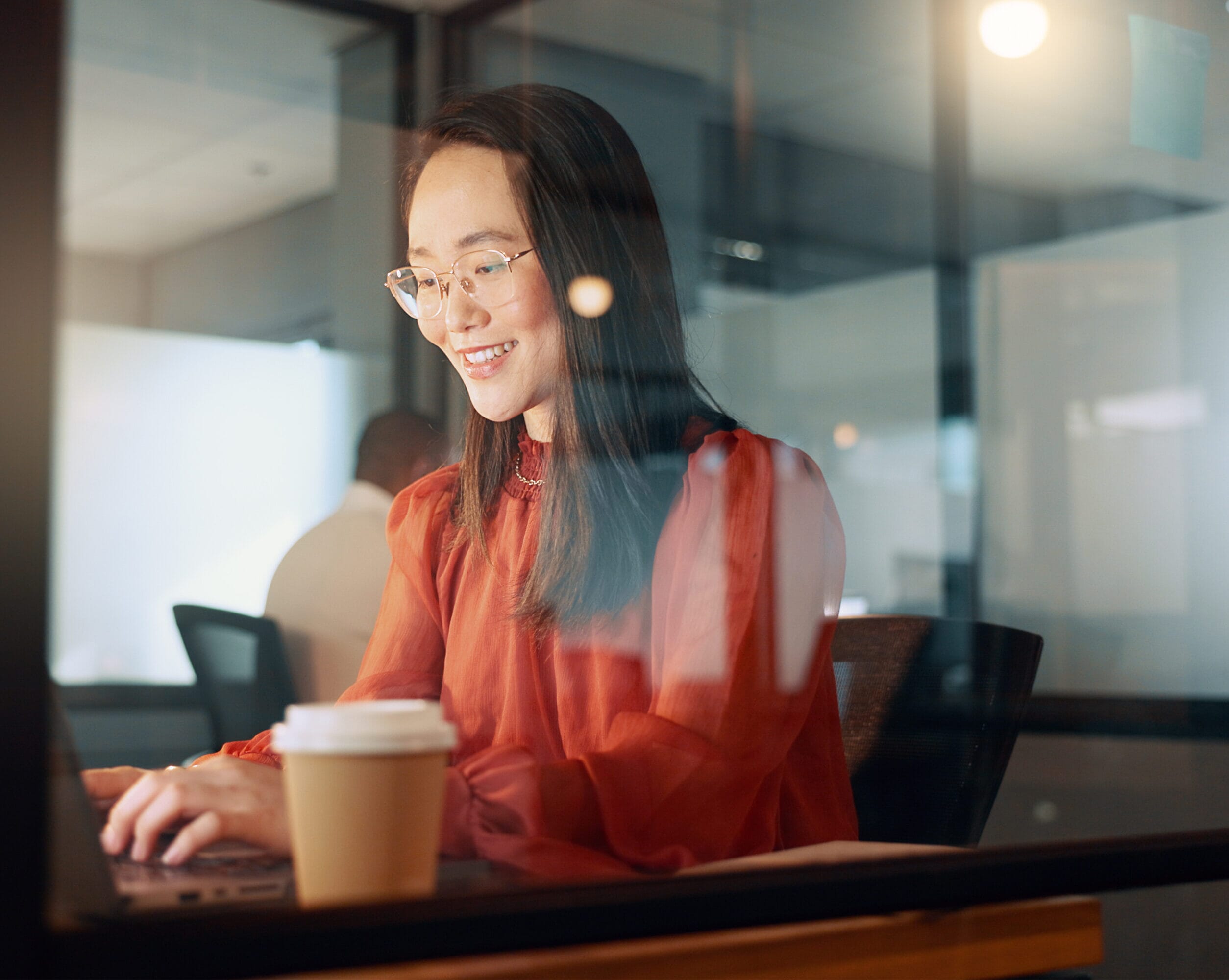 An asian global payroll employee wearing a red blouse, glasses and a necklace. She's typing away on a laptop with a smile in an office setting