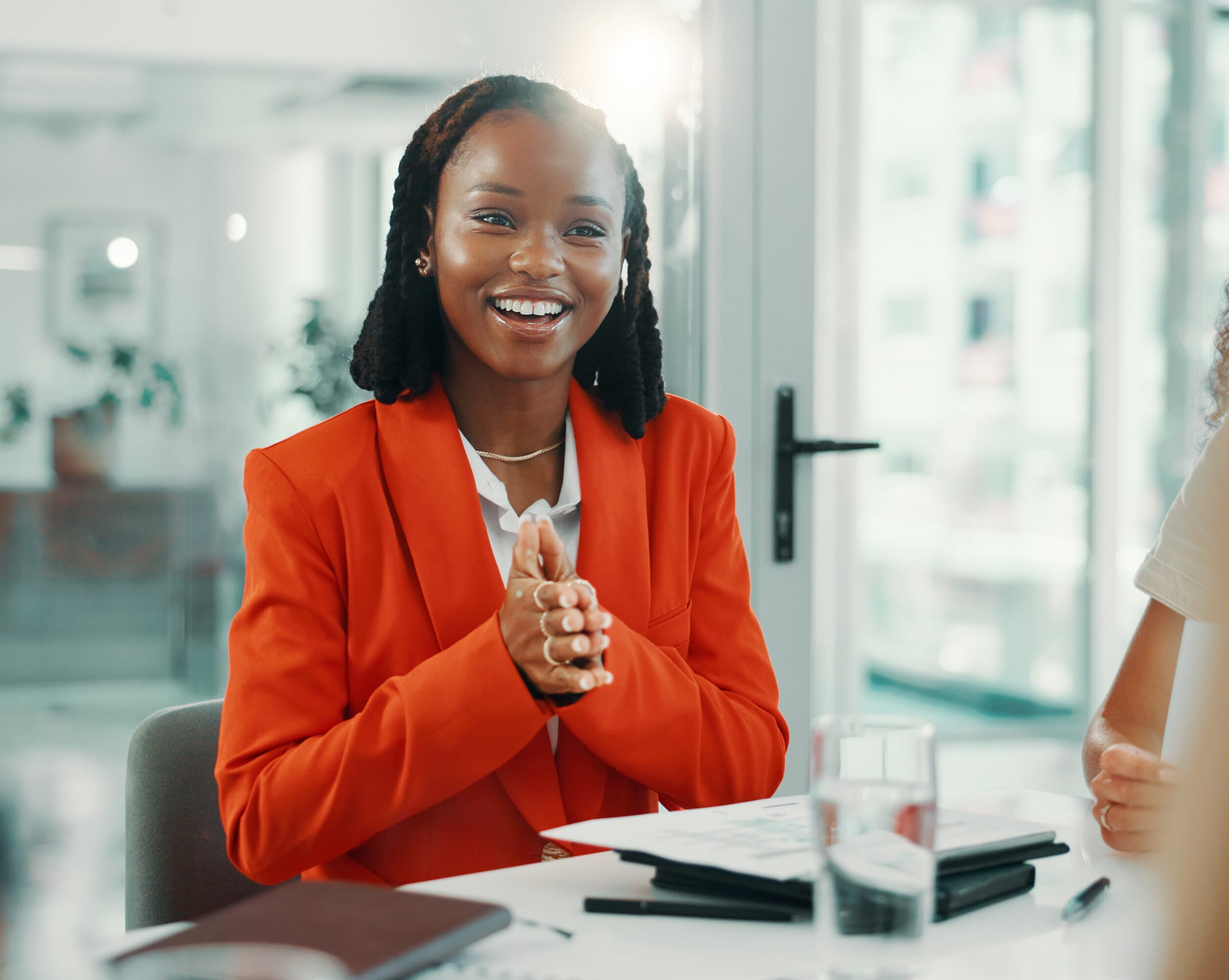 This image goes perfectly with our HR support service offering. A smiling woman in an orange blazer claps her hands while sitting at a conference table, engaging with colleagues in a bright office setting. Glasses of water and notebooks are visible on the table. Choosing outsourced HR services saves you time and money. From our extensive range of people management solutions, we will create a bespoke package of integrated services to simplify processes for attracting, retaining, motivating and supporting employees, all while aligning with corporate goals and meeting legal obligations.
