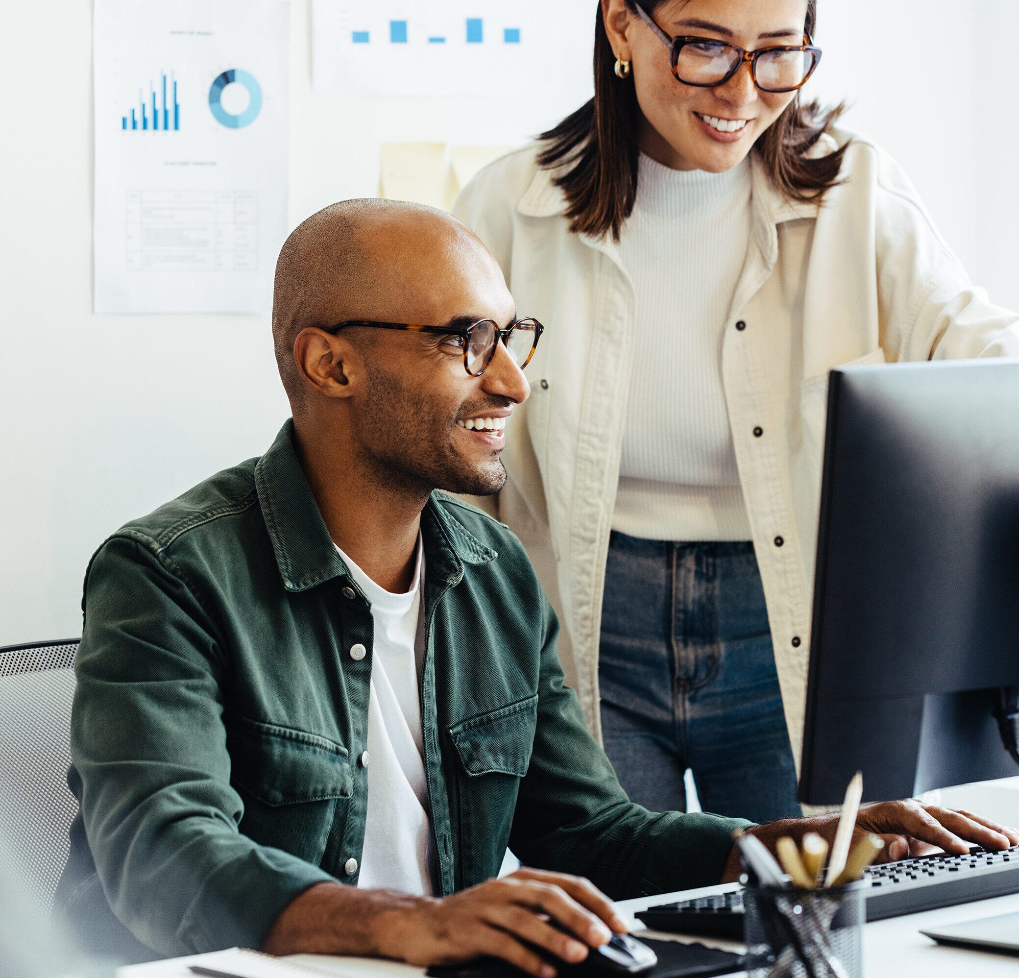 Two colleagues working on an award-winning Payroll Technology client. The man is sat at the desk smiling whilst working on his computer and a lady stood to the left of him also smiling at what he is showing her on the computer.