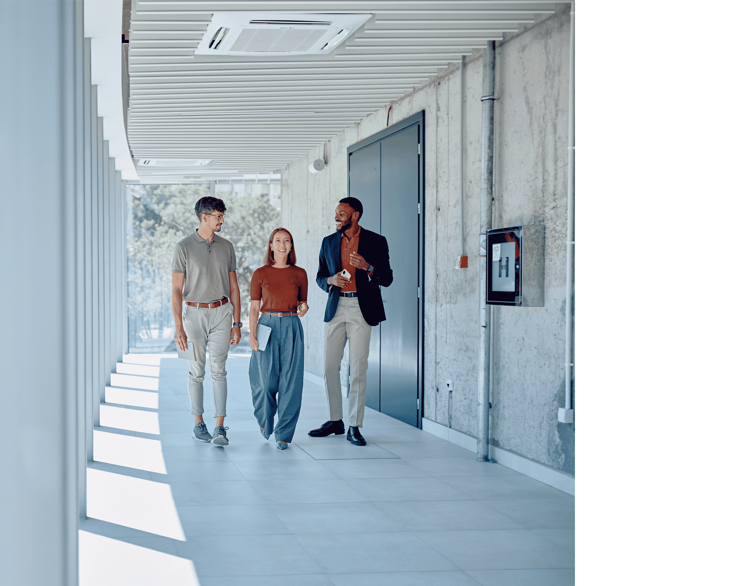3 coworkers walking down a sunlit hallway past an elevator talking about Global staffing. All smartly dressed, the woman walking in between two men is wearing jeans and a red top. The man on the left is wearing smart beige trousers with a darker top. The man on the right is wearing smart white trousers, an orange jumper and a smart navy blazer.