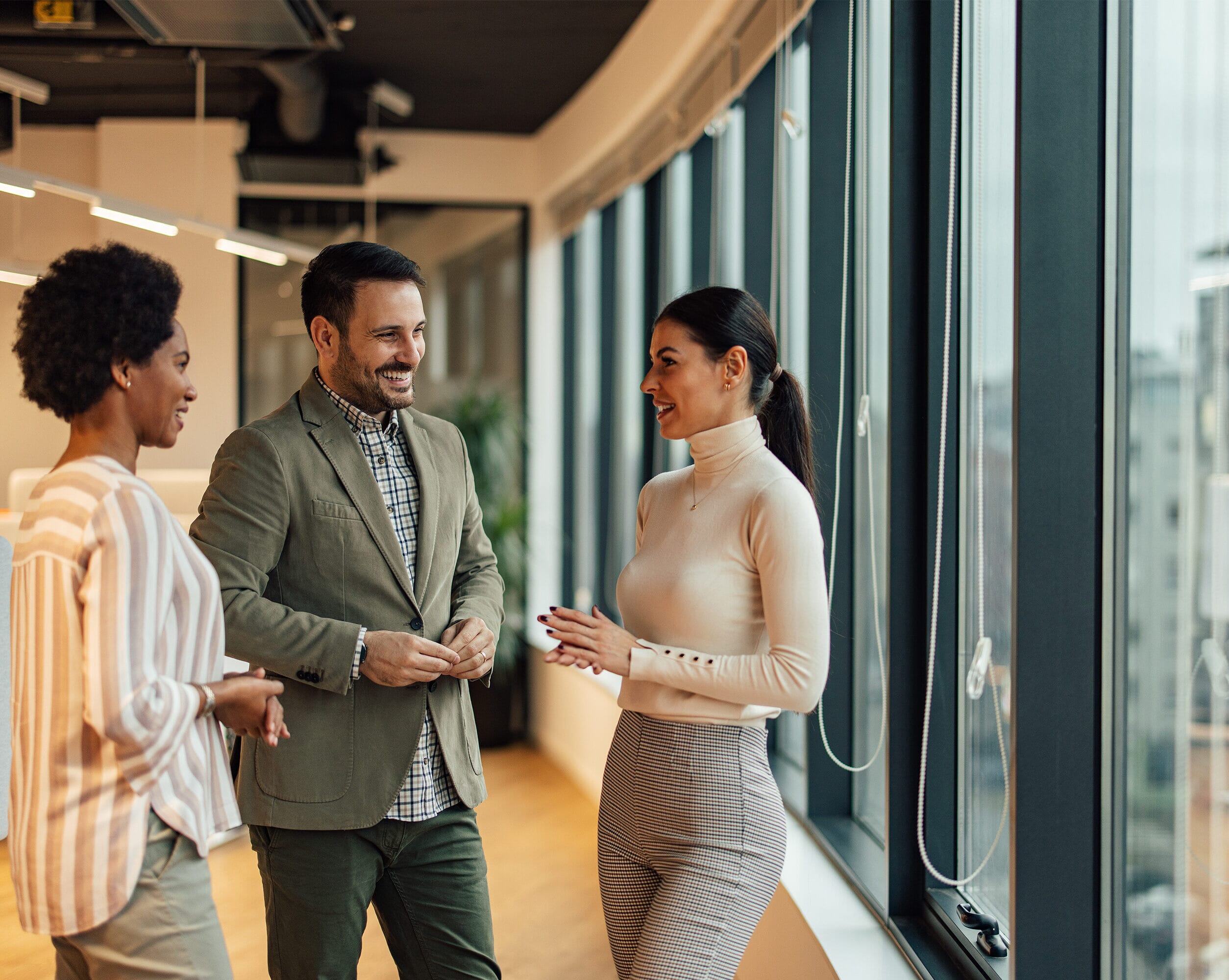 In this image, there are 3 diverse people talking in a hallway of an office building. The Americas present a wide spectrum of growth opportunities across both established and emerging markets. With regional expertise and cross-border capabilities, we deliver customised outsourcing solutions that optimise operations, increase efficiency, and support long-term success throughout North, Central, and South America.
